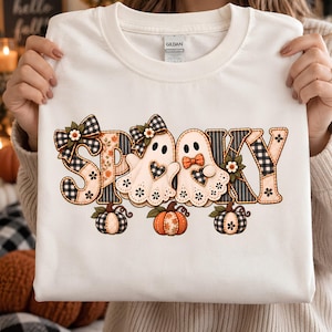 May include: A white t-shirt featuring the word SPOOKY in a decorative font. The letters are filled with black and white buffalo plaid, floral, and striped patterns. Two ghosts with bows and hearts, plus three pumpkins, are integrated into the design.