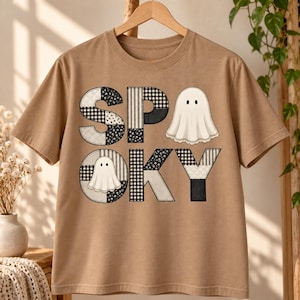 Op de afbeelding: Een beige t-shirt met korte mouwen en het woord SPOOKY in een patchwork-stijl lettertype. De letters zijn gevuld met zwart-wit ruitjespatronen, bloemmotieven en effen vlakken. Twee witte spookillustraties zijn in de letters verwerkt.