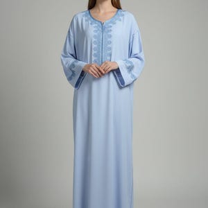 Peut inclure: Un caftan bleu clair pleine longueur avec des manches longues et une encolure ronde. Le vêtement est orné de broderies ton sur ton bleu complexe autour du col, le long de la patte de boutonnage centrale et sur les poignets des manches.