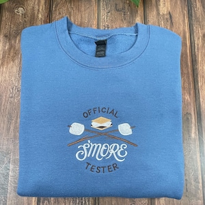 May include: A light blue sweatshirt with the embroidered text "OFFICIAL SMORE TESTER". The design includes a s'more between two marshmallows on sticks. The sweatshirt is folded on a wooden surface.