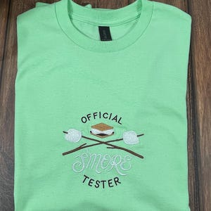 May include: A light green t-shirt with the embroidered text "OFFICIAL SMORE TESTER" and a graphic of a s'more with marshmallows on sticks. The design is in white, brown, and tan.
