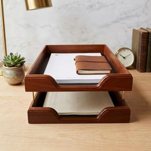 May include: A two-tiered wooden document tray with a rich brown finish and gold-toned metal supports. The top tray holds a stack of white paper and a small brown leather-bound notebook, while the bottom tray contains a stack of white paper.