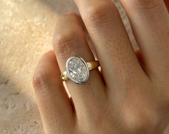 May include: A ring featuring a clear, oval-cut gemstone set in a smooth, silver-toned bezel. The band is crafted from a polished gold-toned metal, creating a two-tone appearance that highlights the central stone's faceted surface and clarity.
