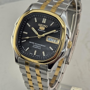 May include: A Seiko 5 Superior automatic wristwatch with a black textured dial, gold-tone bezel, and a two-tone silver and gold stainless steel link bracelet. The dial features gold hands, hour markers, a day-date window, and text reading SEIKO 5 Superior.