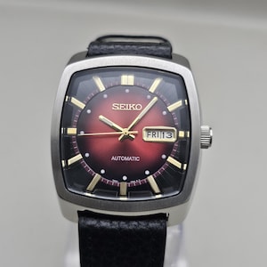 Peut inclure: Une montre-bracelet automatique Seiko avec un boîtier carré argenté et un bracelet en cuir noir. Le cadran présente un dégradé du rouge au noir, des index dorés, des aiguilles dorées et un guichet jour-date à trois heures affichant FRI 13.