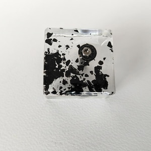Peut inclure: Un bouton carré en résine transparente incrusté de flocons noirs aux formes irrégulières et dentelées. Une base circulaire noire avec un trou de vis central en métal argenté est située sur la surface supérieure du bouton carré.