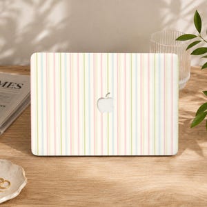 Puede incluir: Una funda para portátil con un patrón de rayas verticales en tonos pastel suaves de rosa, azul claro y verde pálido sobre un fondo crema. El diseño incluye un recorte central que deja ver el logotipo plateado de Apple en la tapa.