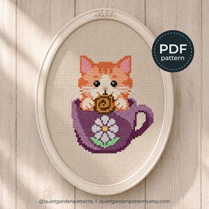 May include: A cross-stitch pattern featuring an orange tabby cat sitting inside a purple teacup decorated with a white daisy. The cat is holding a brown cookie. The design is displayed in an oval frame with a circular PDF pattern label.