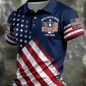 May include: A navy blue polo shirt featuring a graphic of the American flag with white stars on the left and red and white stripes on the bottom. The chest displays an eagle emblem with a cross and the text United States of America 1776-2026.