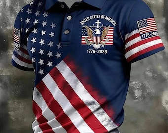 May include: A navy blue polo shirt featuring a graphic of the American flag with white stars on the left and red and white stripes on the bottom. The chest displays an eagle emblem with a cross and the text United States of America 1776-2026.