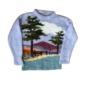 May include: A long-sleeved knit jumper in light blue featuring a landscape scene on the front. The design includes two dark green pine trees, a white cloud, a dark red mountain, a black fence, and a foreground of yellow and teal grass.