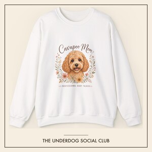 Op de afbeelding: Een wit sweatshirt met lange mouwen en een centrale afbeelding van een bruine Cavapoo-hond omringd door bloemillustraties. De tekst luidt Cavapoo Mom en PROFESSIONAL BABY TALKER. Onderaan de afbeelding staat THE UNDERDOG SOCIAL CLUB.