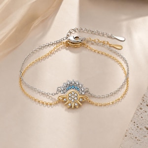 May include: A double-strand bracelet featuring a silver-tone chain with a crescent moon charm and a gold-tone chain with a sun charm. Both charms are embellished with clear crystals, and the moon charm includes small blue stones.