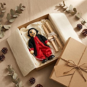 May include: A wooden marionette puppet dressed in a black jacket and red trousers with a black eye patch, resting inside an open cardboard gift box lined with white tissue paper, accompanied by a wooden control bar with white strings attached.