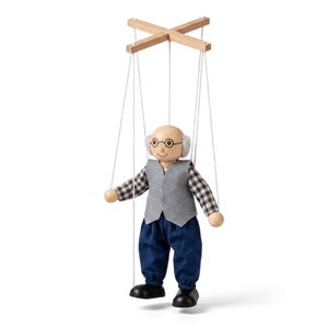 Könnte beinhalten: Eine Holzmarionette einer älteren Person mit Glatze, grauem Haar und runder Brille. Die Figur trägt eine graue Filzweste über einem schwarz-weiß karierten Hemd, eine blaue Hose und schwarze Schuhe und ist an weißen Fäden an einem Holzkreuz aufgehängt.