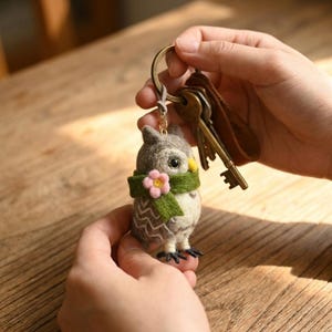 Pode incluir: Um porta-chaves de coruja em lã feltrada com corpo cinzento e branco estampado, um cachecol verde com uma pequena flor rosa e um bico amarelo. A coruja está presa a um anel de metal cor de bronze que segura várias chaves e uma etiqueta de couro castanho.