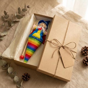 May include: A wizard doll with a blue hat, white beard, and a rainbow-striped cone-shaped body, accompanied by a wooden stick with a red ball tip, presented inside an open rectangular cardboard gift box tied with twine and a blank paper tag.