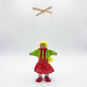 Könnte beinhalten: Eine hölzerne Marionette in Form einer Puppe mit gelben Wollhaaren, einem grünen Oberteil mit roten vertikalen Streifen und einem roten Rock mit weißen Punkten. Die Puppe hängt an weißen Fäden, die an einem hölzernen Steuerkreuz befestigt sind.