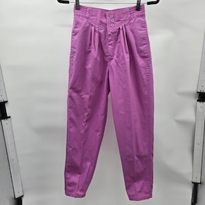May include: High-waisted pants in a solid shade of orchid pink. These trousers feature a structured waistband with three matching buttons, a front zipper closure, and vertical pleats extending from the waist down the front of each leg.