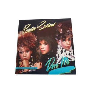 Puede incluir: Una portada de disco de vinilo cuadrada con las Pointer Sisters, con el texto Pointer Sisters en letra cursiva amarilla en la parte superior y Dare Me en letra cursiva turquesa abajo a la derecha. Incluye el texto Special Extended Single en azul.