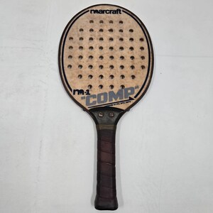 May include: A Marcraft M-1 COMP paddle with a light tan face featuring a grid of circular holes. The paddle has a dark brown handle with a wrapped grip and black text printed on the face, including the brand name and model designation.