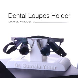 以下が含まれることがあります： Dental Loupes HolderおよびDr. Sumala Yaserと刻印された白いスタンド。グレーのフレーム、黒い円形のレンズ、黒いストラップを備えた歯科用ルーペが置かれています。スタンドの上部にはORGANIZE. WORK. CREATE.という文字があります。