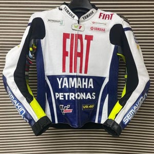 May include: A white, blue, and yellow motorcycle racing jacket featuring the text FIAT, YAMAHA, PETRONAS, and VR 46. The jacket has long sleeves, a front zipper, and various sponsor logos including Packard Bell and WLF printed on the front.