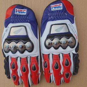 Op de afbeelding: Een paar Honda Racing HRC motorhandschoenen in rood, wit en blauw. De handschoenen hebben metalen knokkelbeschermers met het Honda-logo, HRC-branding en de tekst SCP Concept. De vingers hebben rode lederen accenten en zwarte beschermers.
