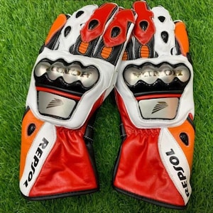 Könnte beinhalten: Ein Paar Repsol Motorradhandschuhe aus rotem, weißem und orangefarbenem Leder. Die Handschuhe verfügen über metallische Knöchelprotektoren mit dem Honda-Logo, schwarze Akzente und den Markennamen Repsol in Schwarz auf den Stulpen.