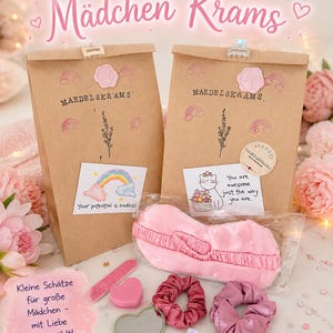 Könnte beinhalten: Zwei braune Papiertüten mit der Aufschrift Maedelskrams, rosa Wachssiegeln, Regenbogen- und Blumenstempeln sowie Karten mit Regenbogen- und Katzenmotiven. Enthalten sind eine rosa Schlafmaske, zwei Stoff-Haargummis, ein herzförmiger Spiegel und eine Nagelfeile.