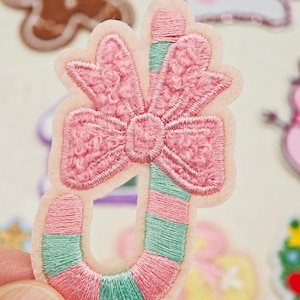 Può includere: Una toppa ricamata a forma di bastoncino di zucchero con strisce diagonali alternate in rosa chiaro e verde menta. La parte superiore del bastoncino è decorata con un grande fiocco rosa testurizzato con un motivo cucito su feltro.