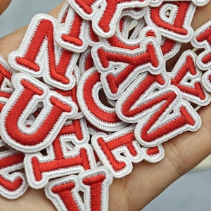 Peut inclure: Une collection de patchs lettres brodés individuels présentant un fil rouge vif pour les caractères et une bordure blanche épaisse. Chaque écusson est en forme de lettre majuscule avec une police à empattement classique pour tissu.