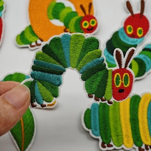 Peut inclure: Un écusson en tissu brodé en forme de chenille verte avec une tête rouge, des yeux jaunes et des antennes brunes. Le corps présente des rayures segmentées dans des tons de vert clair, vert foncé et bleu sarcelle, avec un bord blanc.