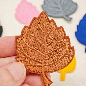Puede incluir: Un parche bordado con forma de hoja marrón con costuras detalladas que muestran una vena central y venas ramificadas. El parche tiene un borde definido y un tallo en la parte inferior. Otros parches de hojas coloridas son visibles.