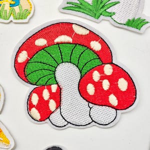 Puede incluir: Un parche bordado que presenta un hongo central con un sombrero rojo cubierto de puntos blancos, una parte inferior verde brillante con láminas visibles y un tallo gris claro. El parche tiene un borde blanco y está rodeado de otros parches de hongos.
