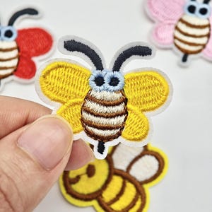 Peut inclure: Un écusson brodé thermocollant en forme d'abeille avec des ailes jaunes, un corps rayé marron et blanc, des yeux bleu clair et des antennes noires. D'autres écussons abeilles et papillons rouges, roses et jaunes sont visibles en arrière-plan.