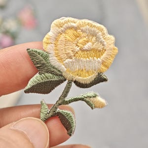 Op de afbeelding: Een geborduurde patch met een gele rozenbloesem met witte accenten, vergezeld van een groene steel, verschillende groene bladeren en een kleine, gesloten gele bloemknop, alles uitgevoerd in gedetailleerd borduurwerk.