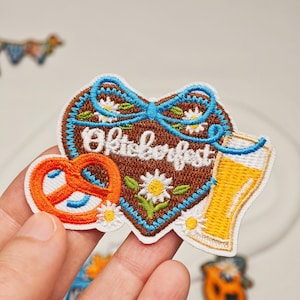 Puede incluir: Un parche bordado con forma de corazón y centro marrón, que presenta el texto blanco Oktoberfest, una cinta azul, margaritas blancas, un pretzel naranja y un vaso de cerveza amarillo, todo bordeado con un contorno blanco sobre una base de tela.