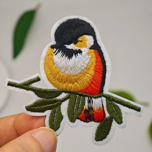Peut inclure: Un écusson brodé représentant un petit oiseau à tête noire, joues blanches, poitrine jaune et plumes d'ailes rouge orangé, perché sur une branche feuillue verte. L'écusson mesure 7,6 par 6,3 centimètres ou 2,99 par 2,48 pouces.