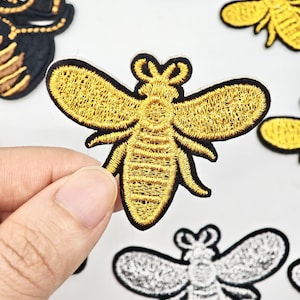Peut inclure: Un écusson brodé en forme d'abeille avec du fil doré métallique pour les ailes et le corps, souligné par une fine bordure noire. Le motif présente des détails de couture sur les ailes et l'abdomen, donnant un aspect texturé à cet insecte.