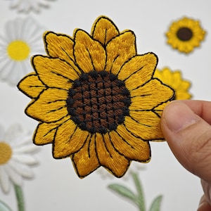 Peut inclure: Un écusson brodé en forme de tournesol avec des pétales jaune vif et un centre texturé brun foncé. L'écusson est présenté devant un fond montrant d'autres écussons brodés de fleurs, notamment des marguerites blanches et un petit tournesol.