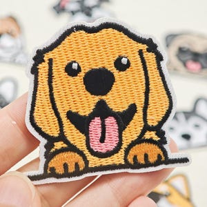 Könnte beinhalten: Ein gestickter Aufnäher mit einem goldenen Cartoon-Hund mit gelbem Fell, einer schwarzen Nase und einer herausgestreckten rosa Zunge. Der Hund ist mit seinen Vorderpfoten auf einer horizontalen Linie dargestellt, in einem detaillierten Stickmuster.