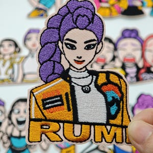 Può includere: Una toppa ricamata raffigurante un personaggio con capelli viola intrecciati, che indossa una giacca gialla sopra una maglietta bianca. Il personaggio ha un'espressione neutra. La parola RUMI è ricamata in grassetto giallo nella parte inferiore.