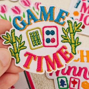 May include: An embroidered patch featuring the text GAME TIME in blue and pink thread. The design includes two mahjong tiles, one with green bamboo sticks and one with four blue and pink dots, flanked by two green bamboo stalks on a white background.