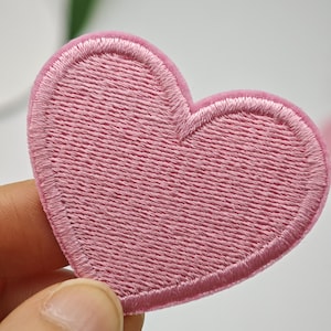 Peut inclure: Un écusson en forme de cœur avec une broderie rose clair présentant un motif de points dense et texturé, ainsi qu'une bordure lisse assortie. Le design est une forme de cœur classique et simple avec un sommet arrondi et une base pointue.