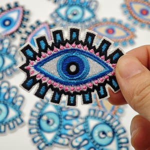 Peut inclure: Un écusson brodé représentant un motif d'œil stylisé avec une pupille noire centrale, un iris bleu et une sclérotique gris clair. L'œil est entouré de détails cousus noirs, bleus et roses, avec un motif de bordure dentelé et rayonnant.