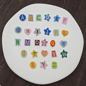 Peut inclure: Une collection de vingt-six écussons brodés représentant les lettres de l'alphabet de A à Z. Les lettres sont présentées dans des formes variées, notamment des cercles, des carrés, des étoiles et des cœurs, avec une palette de couleurs vives.