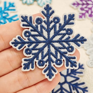 May include: A dark blue embroidered snowflake patch with a white felt backing. The design features intricate geometric branches and circular accents. Additional snowflake patches in light blue, purple, and silver are visible in the background.