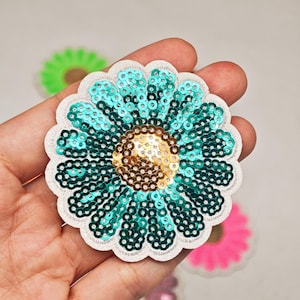 Peut inclure: Un écusson en forme de fleur circulaire avec des paillettes bleu sarcelle sur les pétales et des paillettes dorées au centre. L'écusson présente une bordure brodée blanche et un bord festonné. D'autres fleurs en vert, rose et bleu sont visibles.