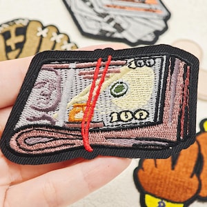 Può includere: Una toppa ricamata raffigurante una mazzetta di banconote da cento dollari fissata con un elastico rosso brillante. La toppa presenta un bordo nero spesso e mostra dettagliate cuciture della valuta, incluso il numero 100 e elementi del ritratto.